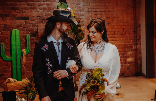 Bride and groom walking away from the alter. Groom is wearing custom embroidered denim shirt with bolo tie and plain white shirt. Holding arms, the couple smile. The bride wears white and holds a bouquet. Set in front of a gothic western dressed venue. 