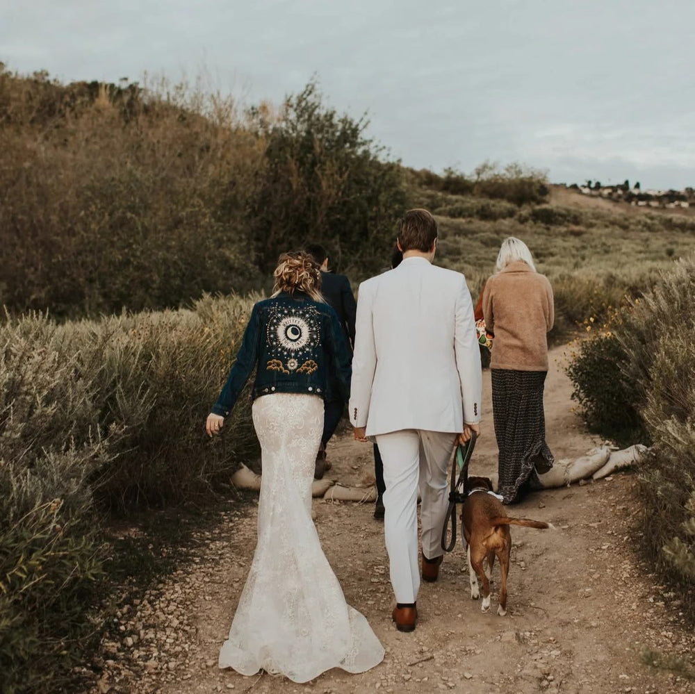 A bride in a white lace mermaid wedding gown and a groom in a white suit walk away on a dirt path with an older woman and a dog, wearing a dark blue denim jacket with gold embroidery on the back.