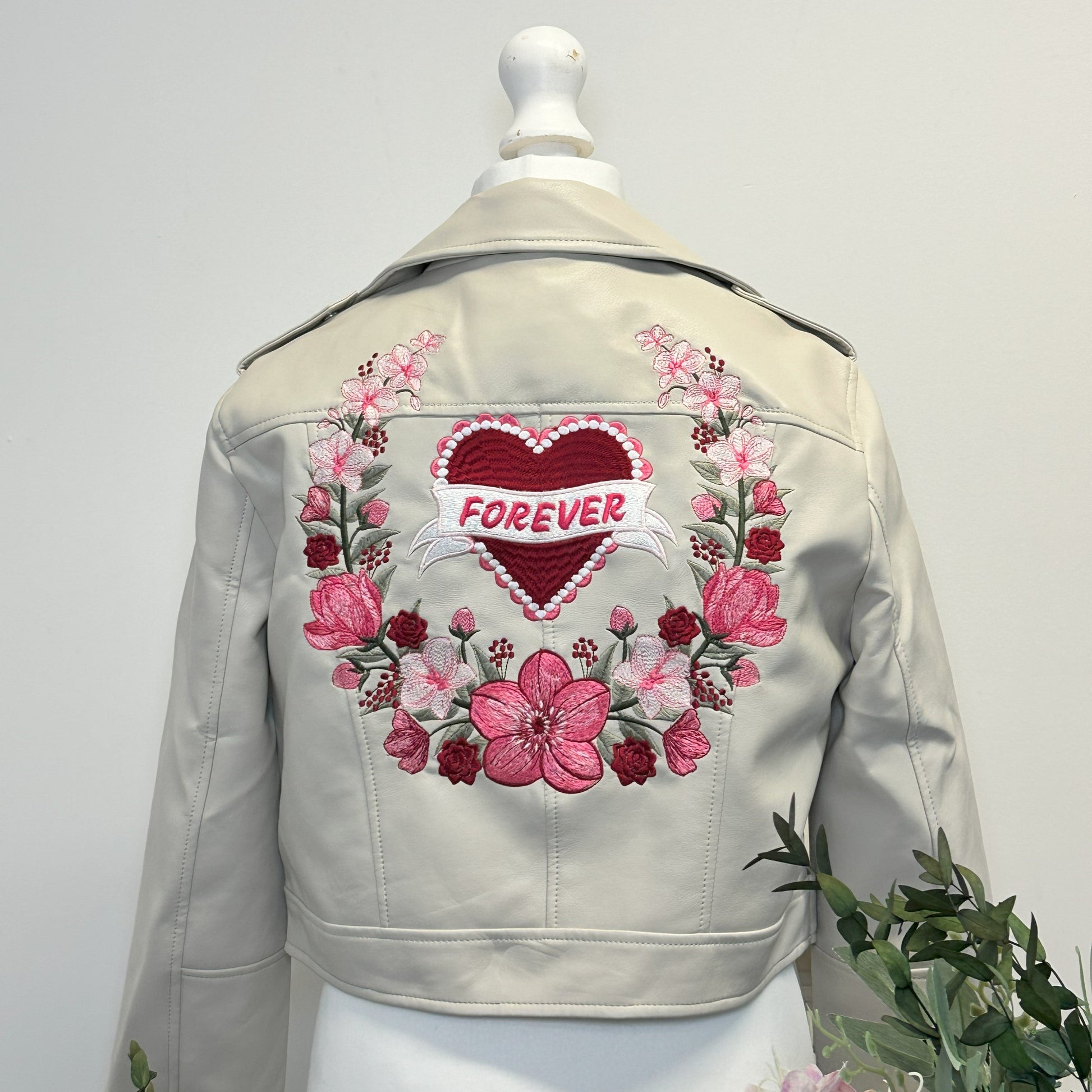 A light beige faux leather jacket displayed on a white mannequin, showing the back decorated with a large pink floral wreath and a central red heart patch with the word 'FOREVER' in white.