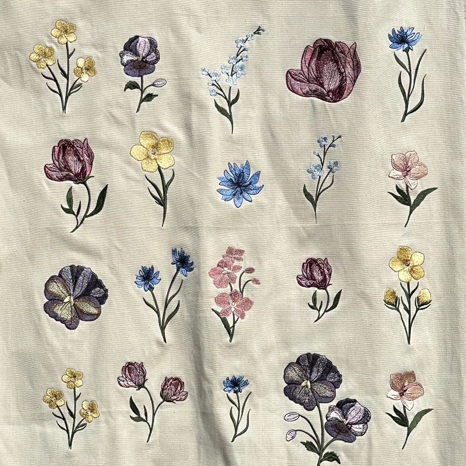 An off-white linen tablecloth with a colorful embroidered floral pattern, hanging from a white metal rack against a red brick wall.