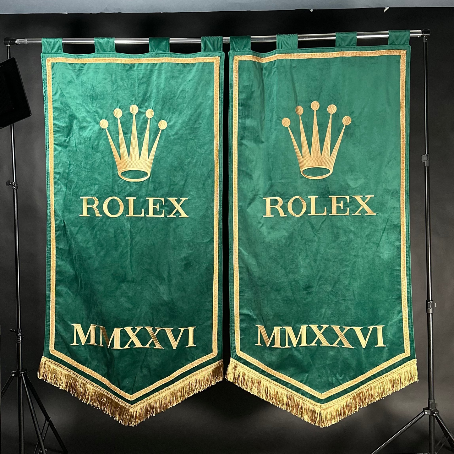 Two green velvet Rolex banners with gold embroidery, hanging from a black metal stand against a dark studio backdrop.