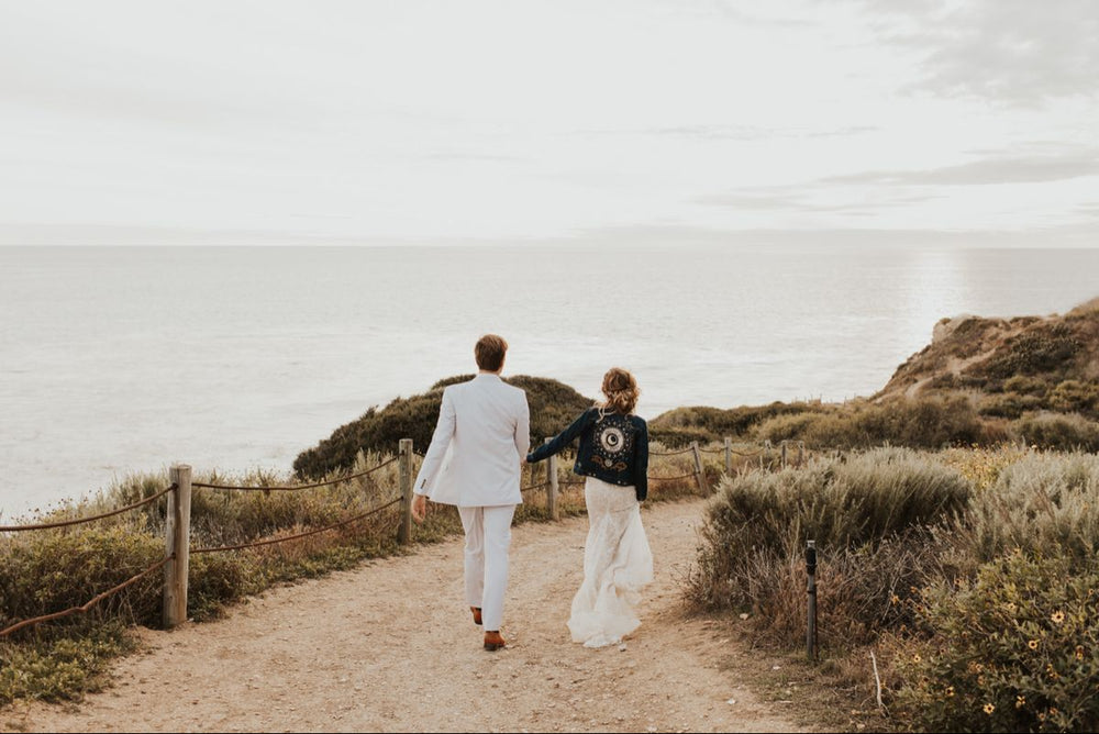 A bride and groom walking away from the camera along a dirt path next to the coast. The bride is wearing a dark indigo denim jacket with gold embroidery on the back. 