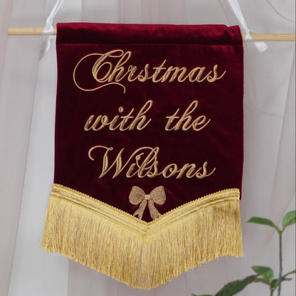 A personalized Christmas banner made of deep red velvet with gold embroidered text reading 'Christmas with the Wilsons' and a gold bow, hanging from a wooden dowel with white hanging ribbons.
