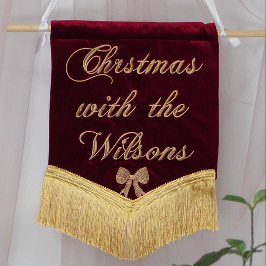 A personalized Christmas banner made of deep red velvet with gold embroidered text reading 'Christmas with the Wilsons' and a gold bow, hanging from a wooden dowel with white hanging ribbons.