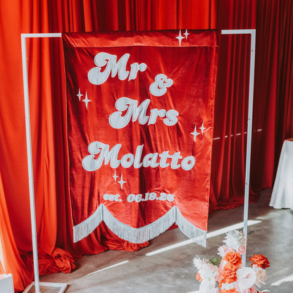 A red velvet wedding backdrop banner on a white metal stand, featuring the text 'Mr & Mrs Molatto' in white script with decorative sparkles and a white fringe hem. The banner is set against a background of red curtains, with a floral arrangement on the concrete floor in the foreground.