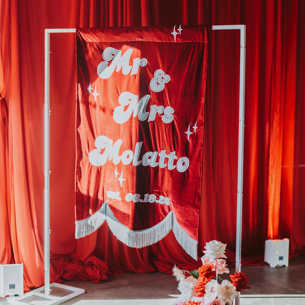 A red velvet wedding banner with white script reading 'Mr & Mrs Molatto' and a white fringe hem, displayed on a white metal stand in front of a red curtain backdrop.