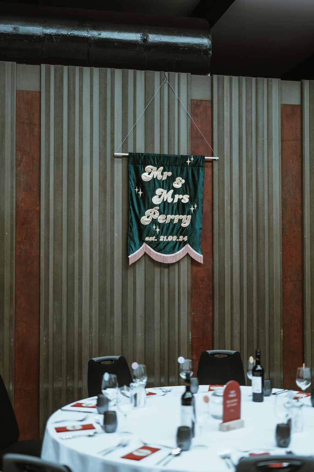 A luxury embroidered banner with tassel on the back wall of a wedding reception room, embroidered with "Mr & Mrs Perry" + wedding date. 