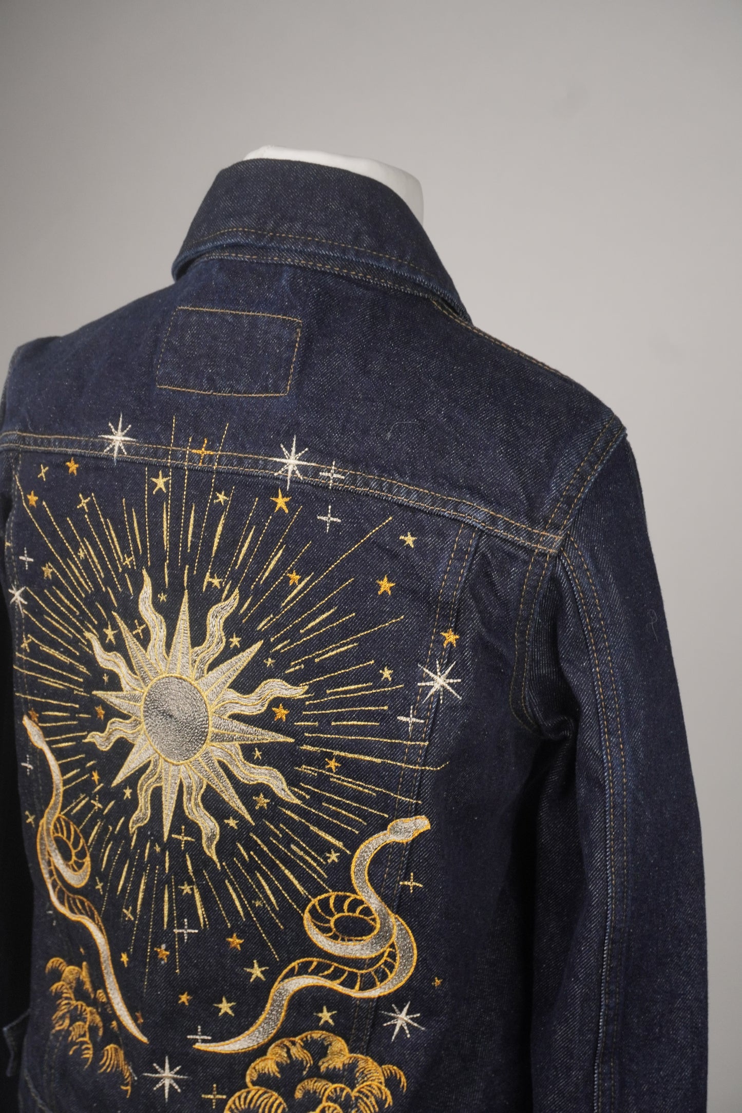 A dark blue denim jacket shown from the back on a mannequin, featuring a large celestial sun and snake embroidery in gold and white thread.