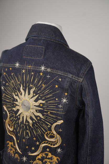 A dark blue denim jacket shown from the back on a mannequin, featuring a large celestial sun and snake embroidery in gold and white thread.