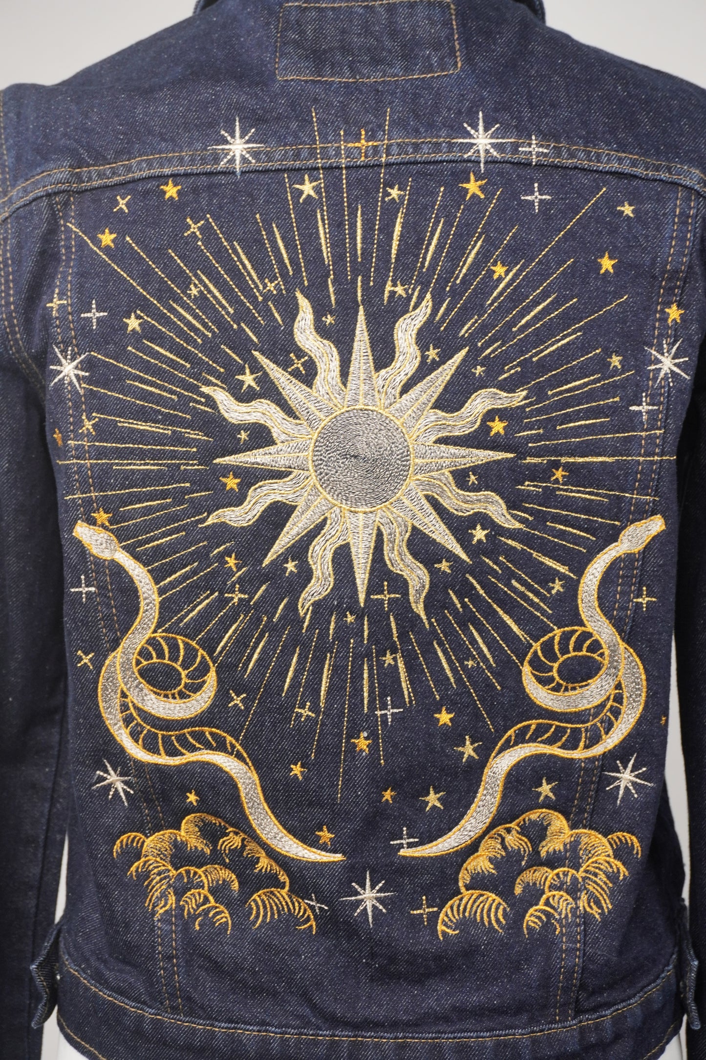 Close-up of a dark blue denim bag featuring intricate gold and white embroidered celestial designs, including a central sun, surrounding stars, and two coiled snakes.