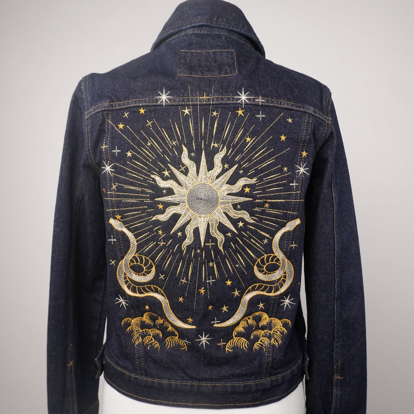 A dark blue denim jacket displayed on a white mannequin torso, showing the back panel decorated with a large celestial sunburst design in gold and silver thread with stars and clouds.