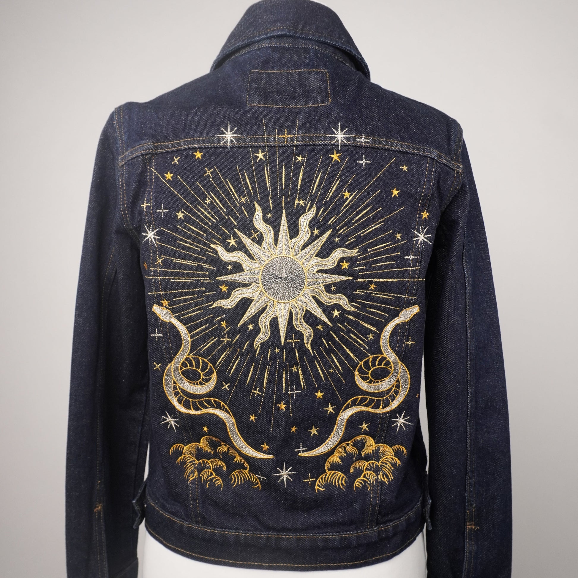 A dark blue denim jacket displayed on a white mannequin torso, showing the back panel decorated with a large celestial sunburst design in gold and silver thread with stars and clouds.