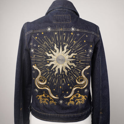 A dark blue denim jacket displayed on a white mannequin torso, showing the back panel decorated with a large celestial sunburst design in gold and silver thread with stars and clouds.