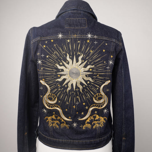 A dark blue denim jacket displayed on a white mannequin torso, showing the back panel decorated with a large celestial sunburst design in gold and silver thread with stars and clouds.