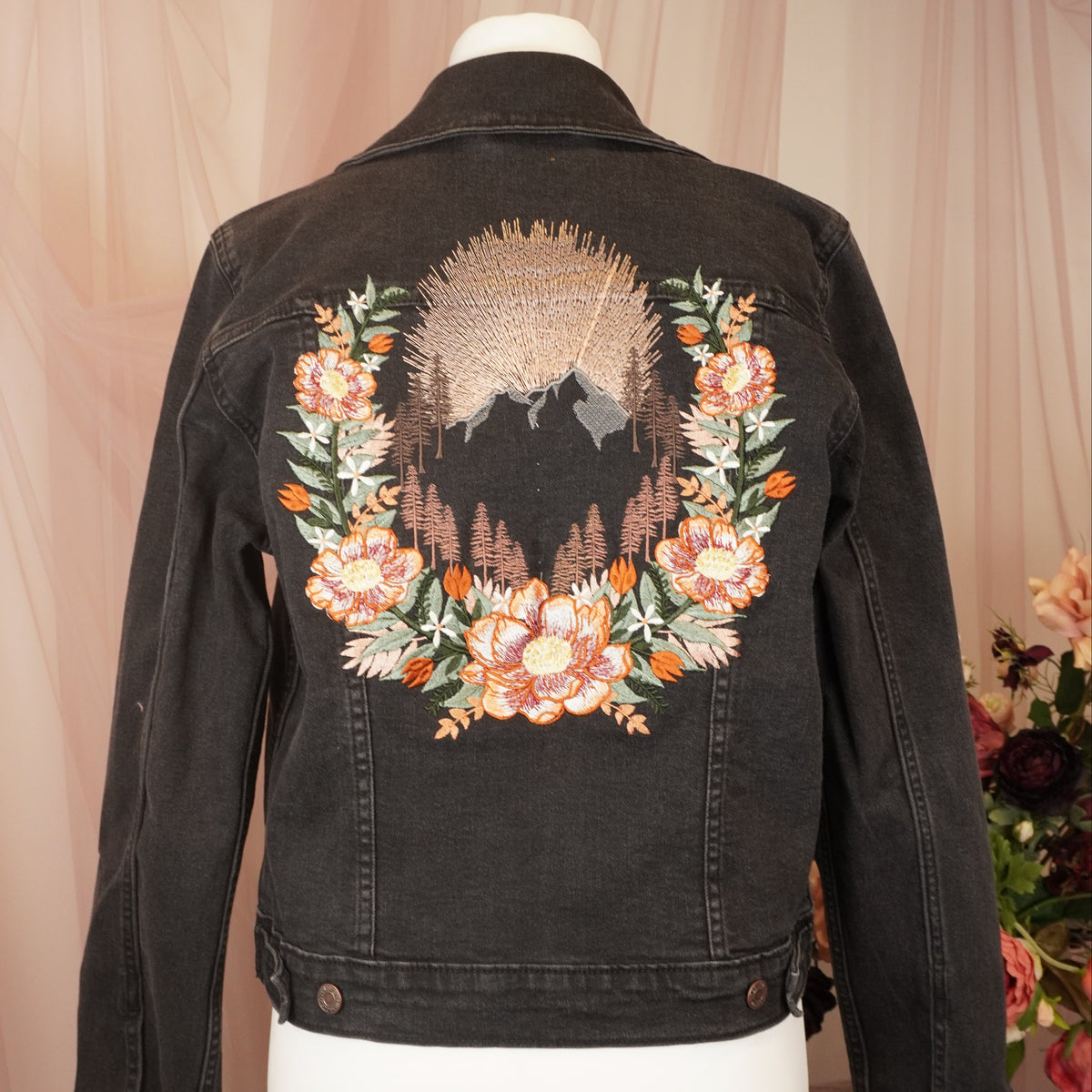 A black denim jacket displayed on a white mannequin torso, featuring a large embroidered design on the back with a floral wreath and a mountain landscape motif in peach, green, and brown tones.