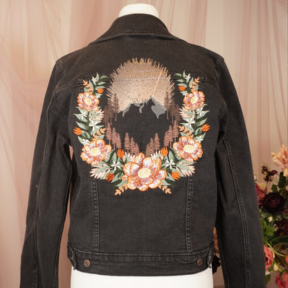 A black denim jacket displayed on a white mannequin torso, featuring a large embroidered design on the back with a floral wreath and a mountain landscape motif in peach, green, and brown tones.