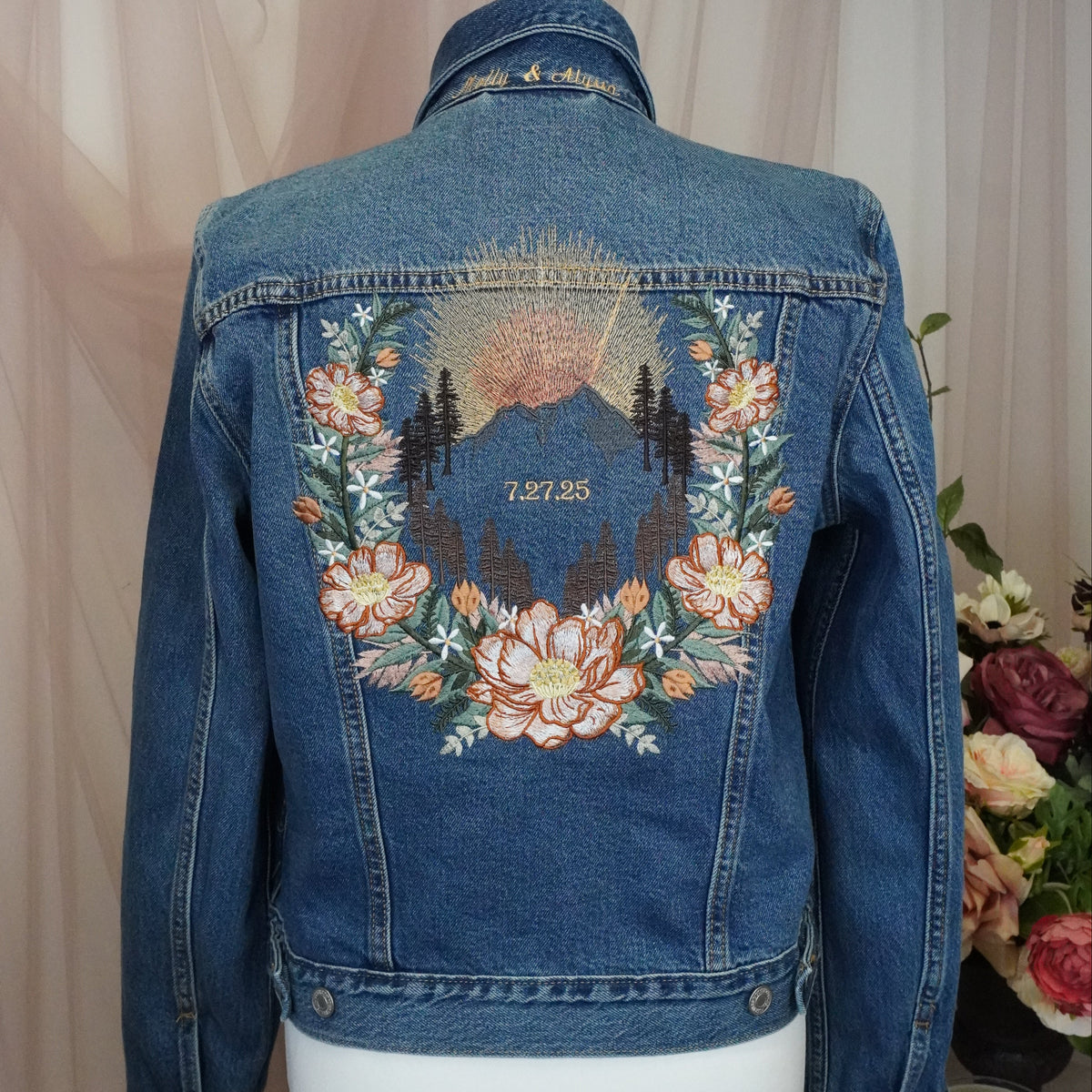 A blue denim jacket displayed on a white mannequin torso, showing the back with a large embroidered floral and mountain design in peach, pink, green, and gold tones. The jacket is photographed against a soft beige draped fabric backdrop with flowers and a candle visible at the side.