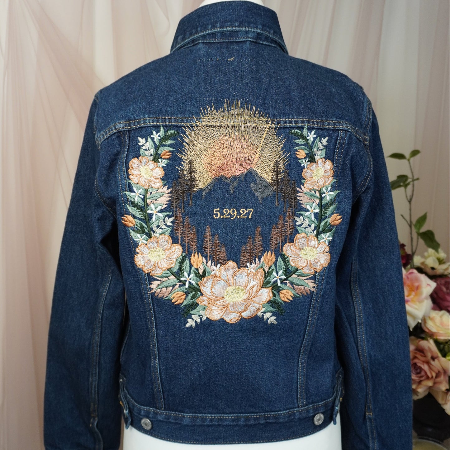 A dark blue denim jacket displayed on a white mannequin torso, showing the back decorated with a large embroidered floral wreath and mountain design in peach, green, and gold tones.