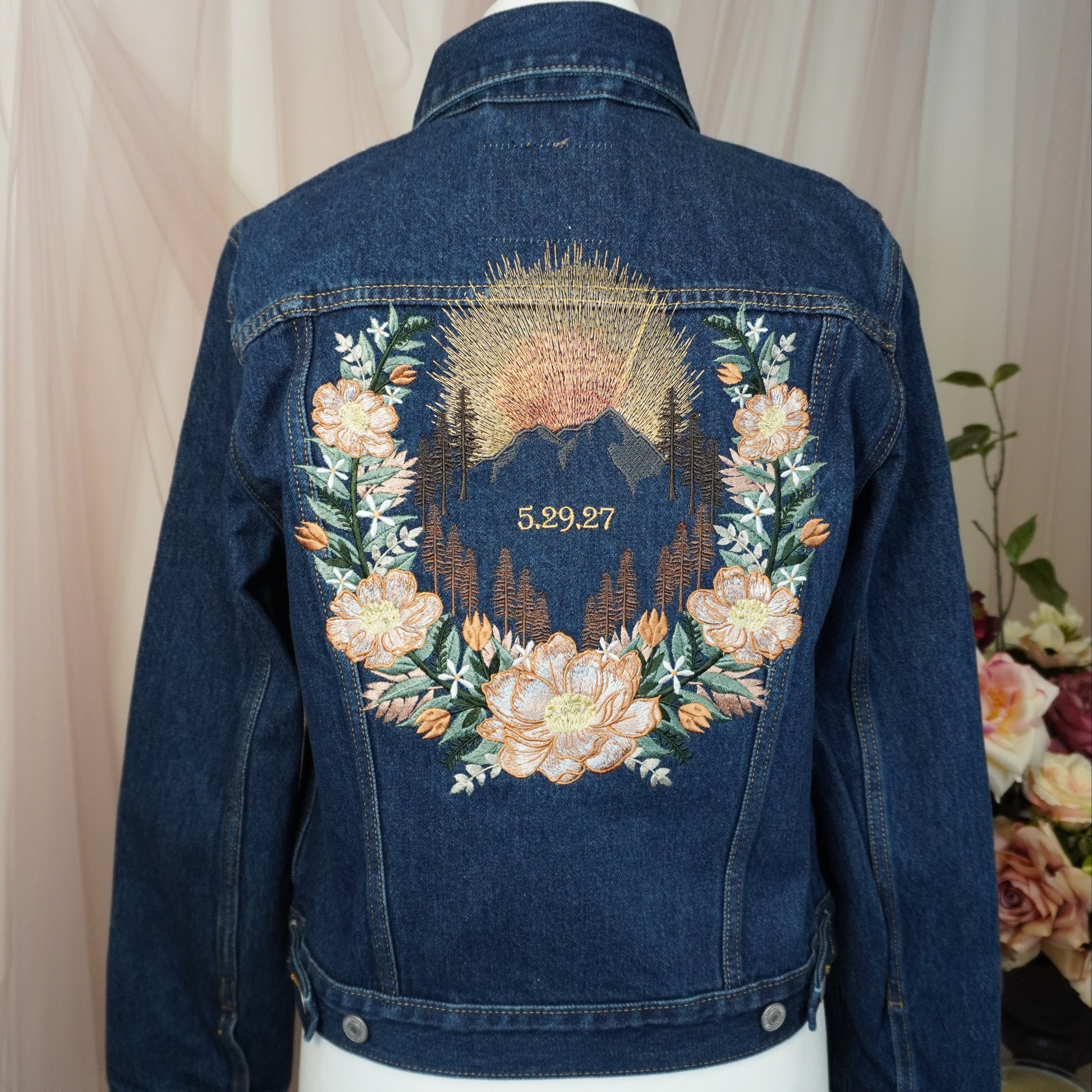 A dark blue denim jacket displayed on a white mannequin torso, showing the back decorated with a large embroidered floral wreath and mountain design in peach, green, and gold tones.