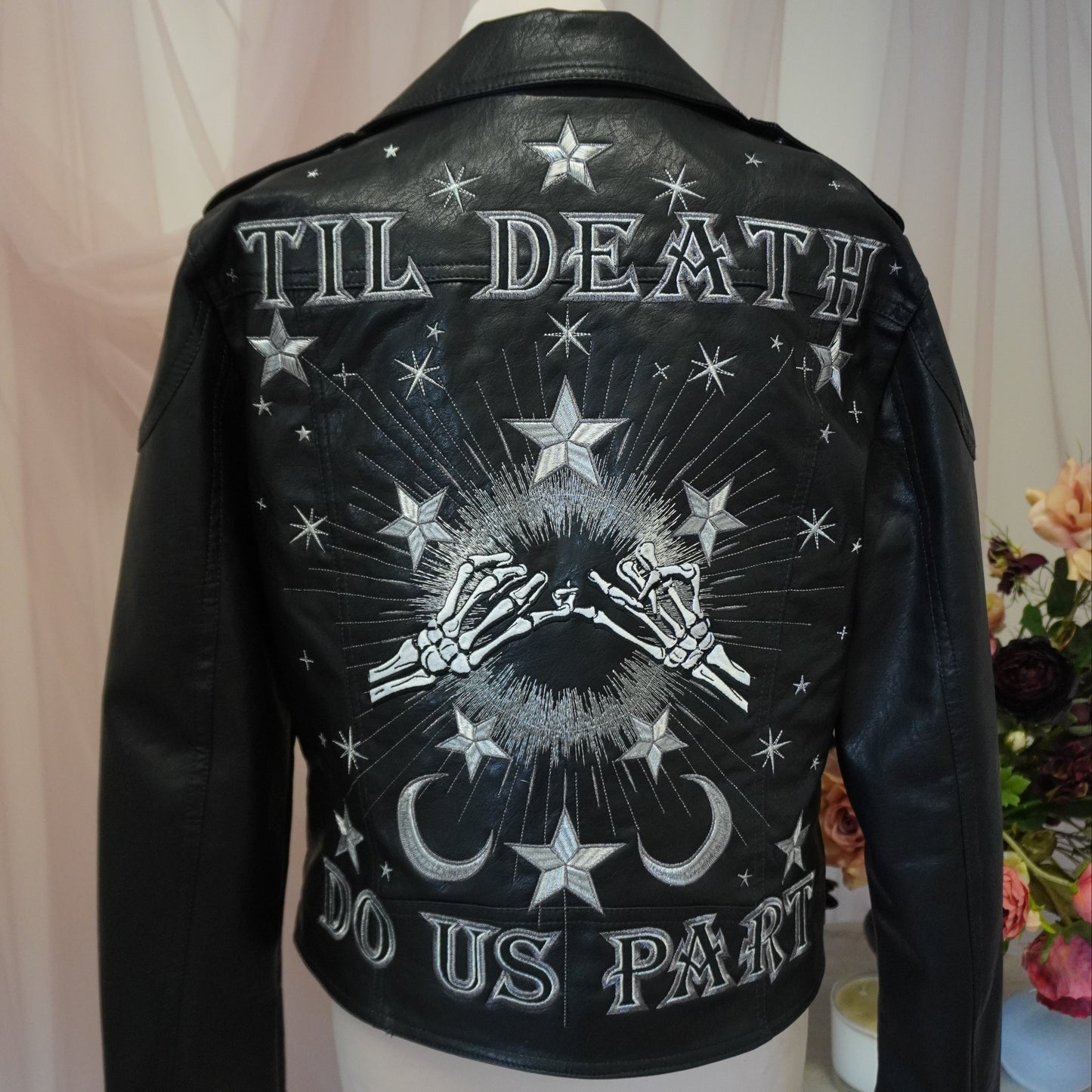 A black leather jacket displayed on a mannequin torso, showing the back with a large white graphic print featuring stars, skeletal hands, a crescent moon, and the text 'TIL DEATH DO US PART'.