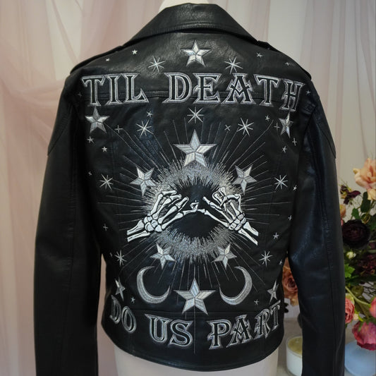 A black leather jacket displayed on a mannequin torso, showing the back with a large white graphic print featuring stars, skeletal hands, a crescent moon, and the text 'TIL DEATH DO US PART'.
