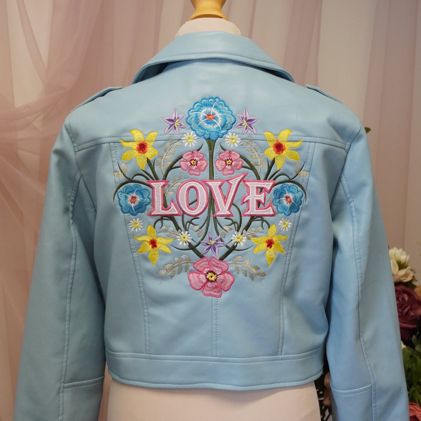 A light blue cropped faux-leather jacket displayed on a cream dress form mannequin, showing a colorful floral and 'LOVE' embroidered design on the back.