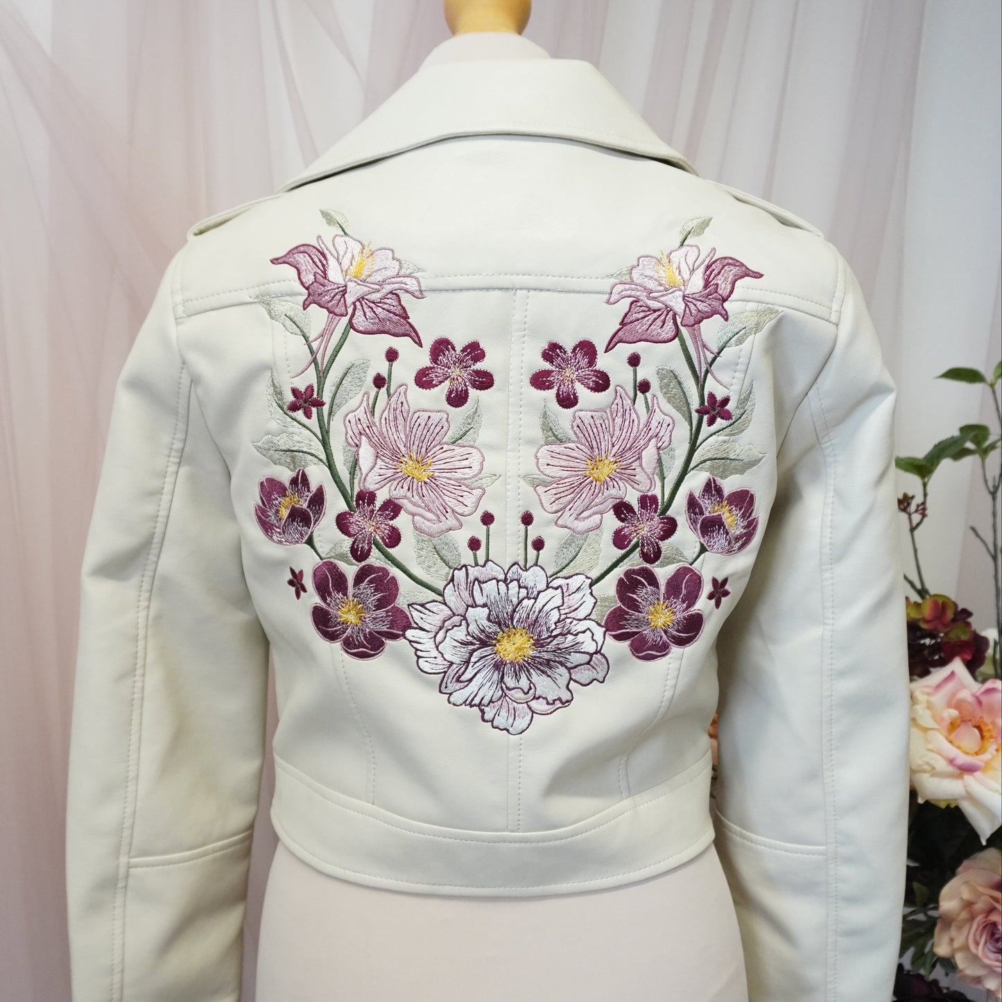 An off-white jacket or cardigan displayed on a cream mannequin torso, featuring a prominent embroidered floral pattern in purple, pink, and white on the back.