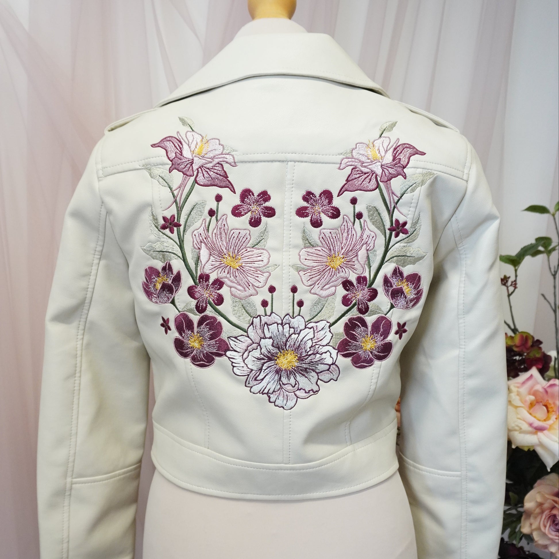 An off-white jacket or cardigan displayed on a cream mannequin torso, featuring a prominent embroidered floral pattern in purple, pink, and white on the back.