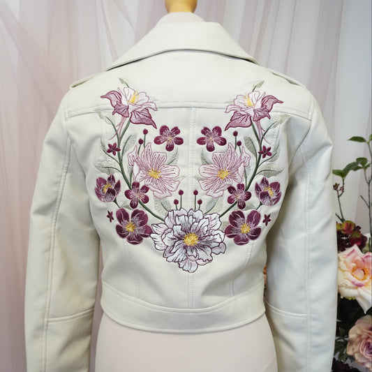 An off-white jacket or cardigan displayed on a cream mannequin torso, featuring a prominent embroidered floral pattern in purple, pink, and white on the back.