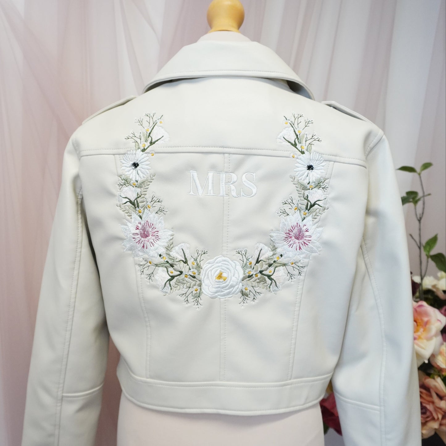 An off-white faux leather jacket displayed on a cream mannequin torso, showing the back decorated with a floral embroidered wreath and the word 'MRS' in white.