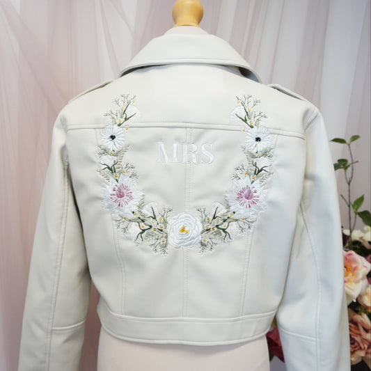 An off-white faux leather jacket displayed on a cream mannequin torso, showing the back decorated with a floral embroidered wreath and the word 'MRS' in white.