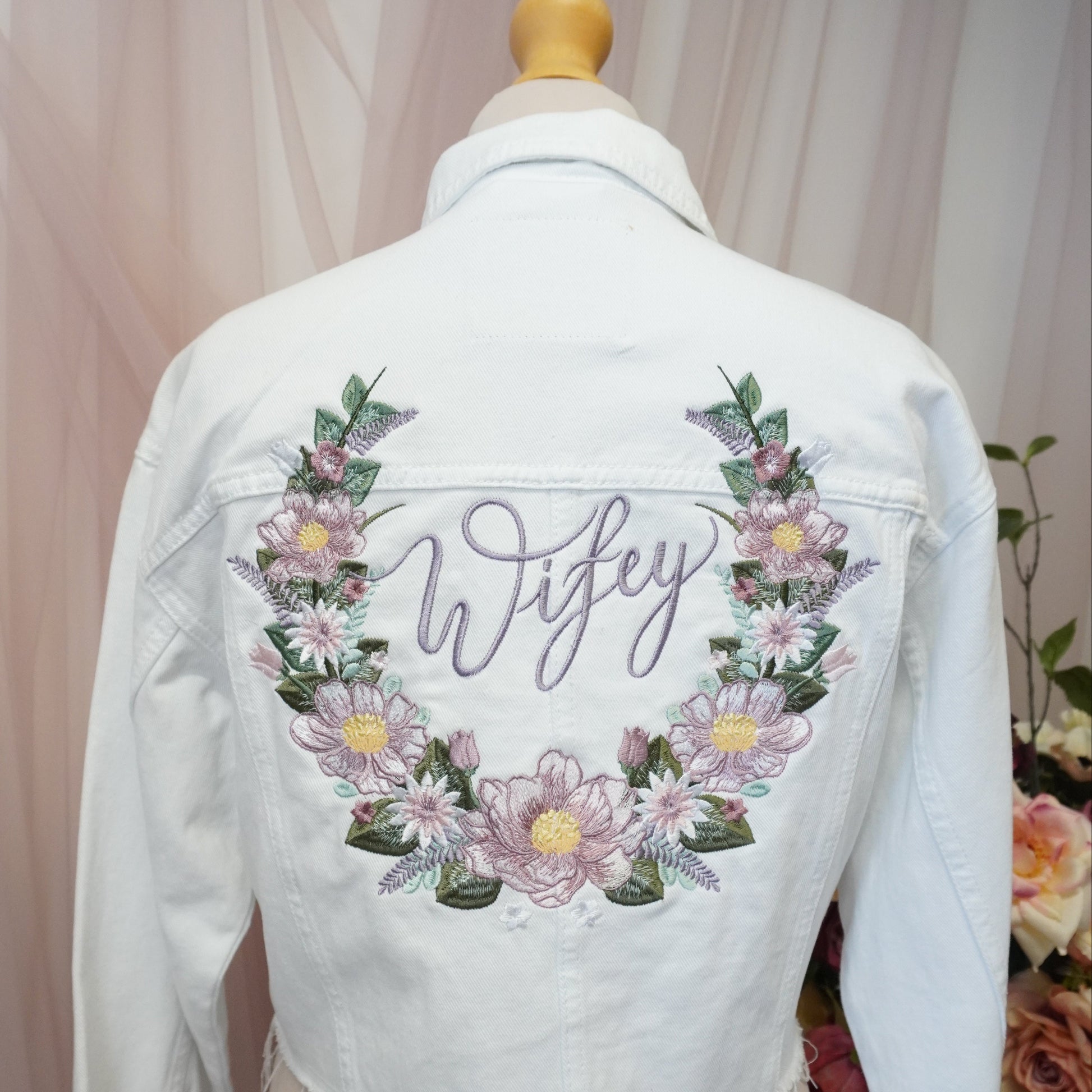 A white denim jacket displayed on a mannequin from the back, featuring a floral wreath design with the word 'Wifey' embroidered in purple script in the center.