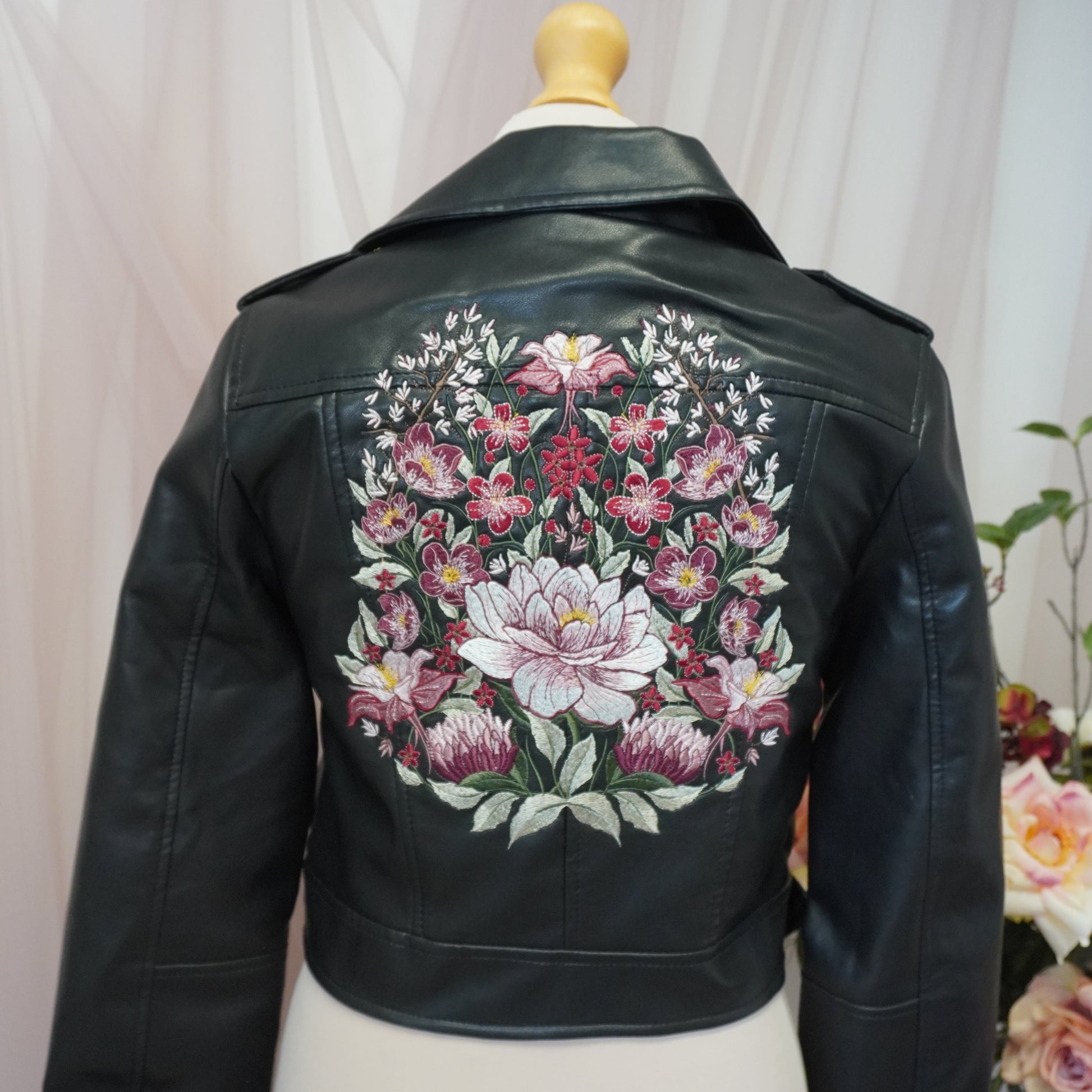 Black leather jacket displayed on a cream dress form, showing a large, colorful floral embroidery design on the back.