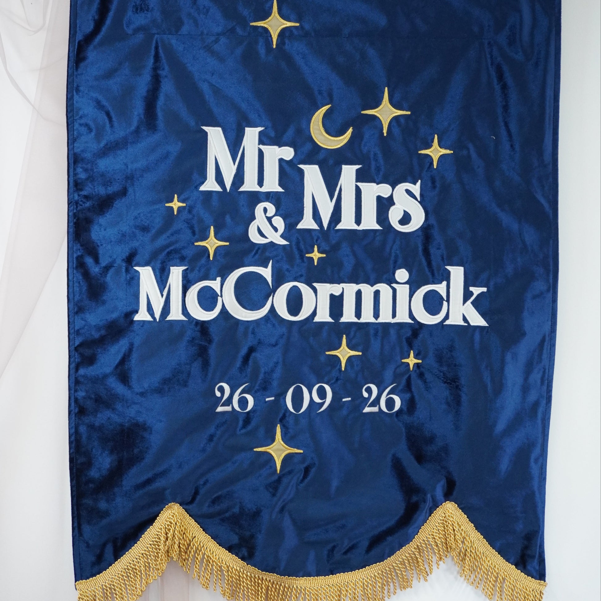 A navy blue satin hanging banner with gold fringe, featuring the text 'Mr & Mrs McCormick' and the date '26-09-26' in white, decorated with gold stars and a crescent moon.