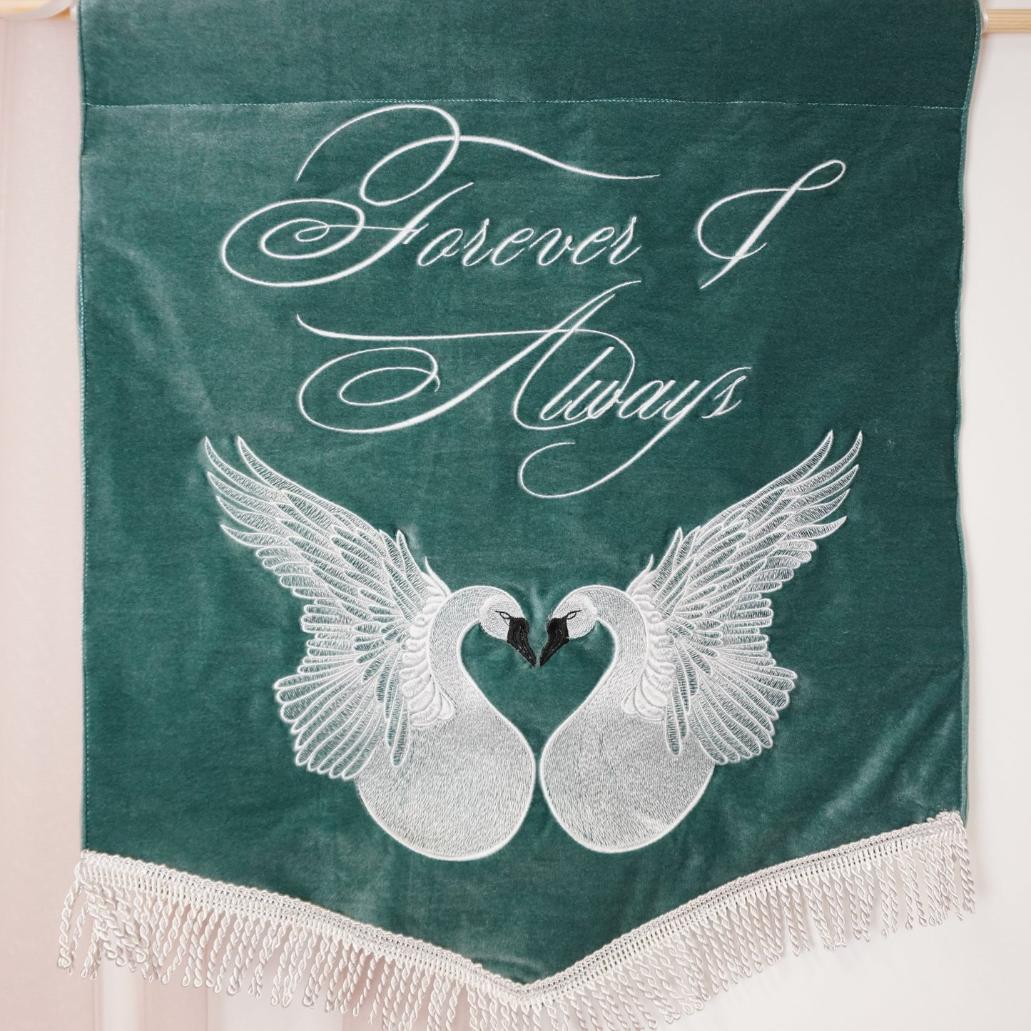 A dark green velvet hanging banner with white embroidered script reading 'Forever & Always' above two white swans facing each other, finished with fringe along the bottom and hanging from a wooden dowel.