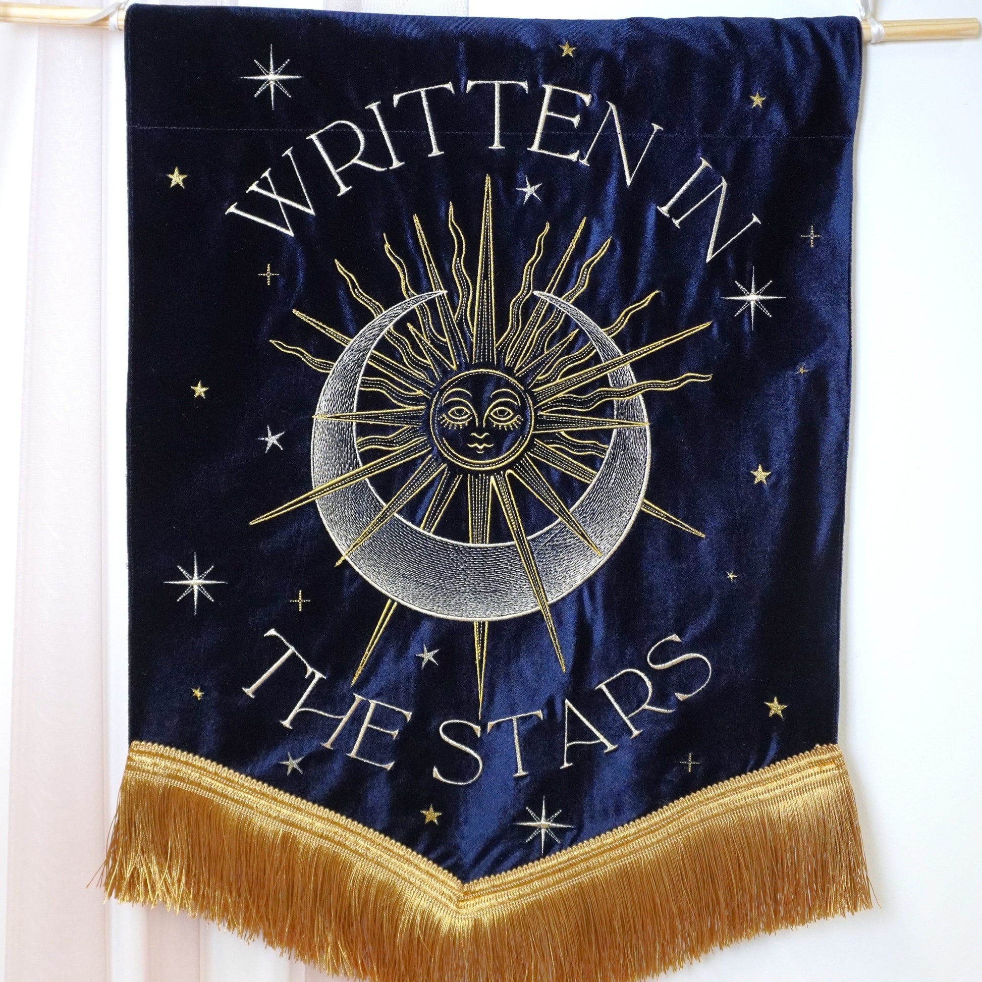 A navy blue velvet wall banner with gold fringe, featuring a gold sun and moon design with the text 'WRITTEN IN THE STARS' and small gold stars, hanging from a wooden dowel against a white curtain background.