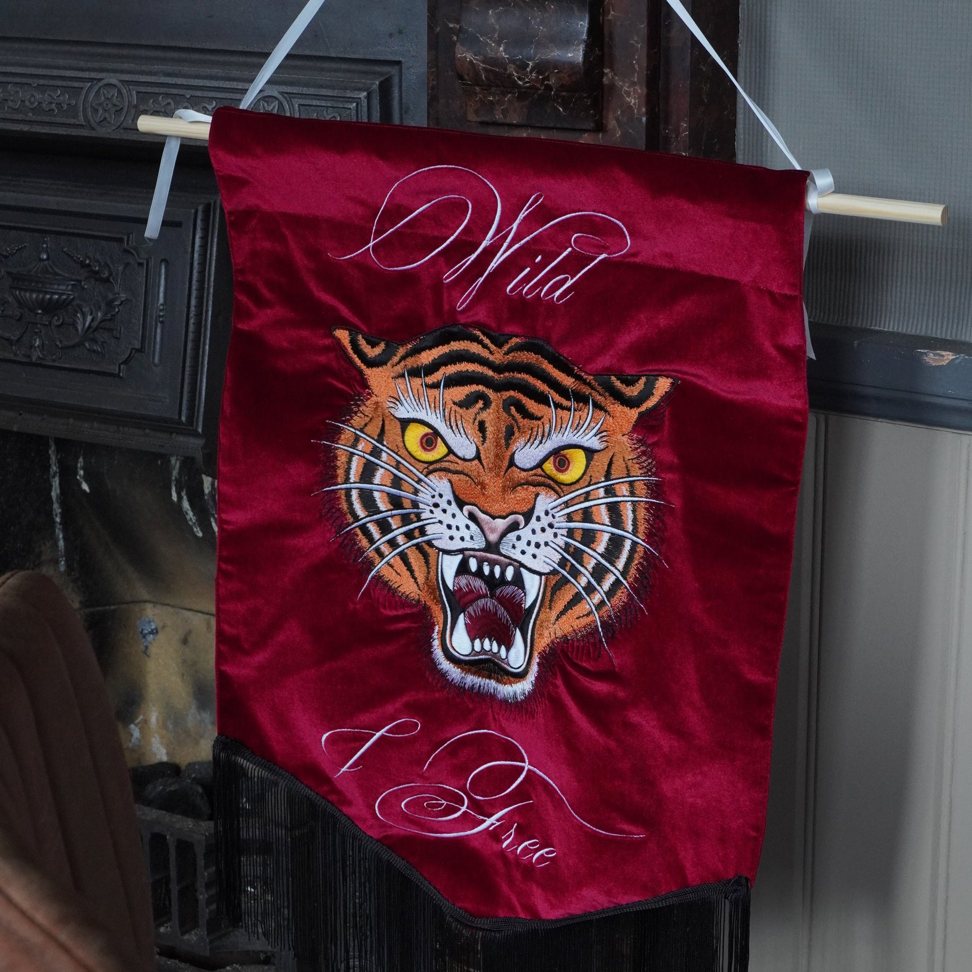 A red velvet banner featuring a roaring tiger face and the words 'Wild & Free' in white script, hanging from a wooden dowel in an indoor setting.