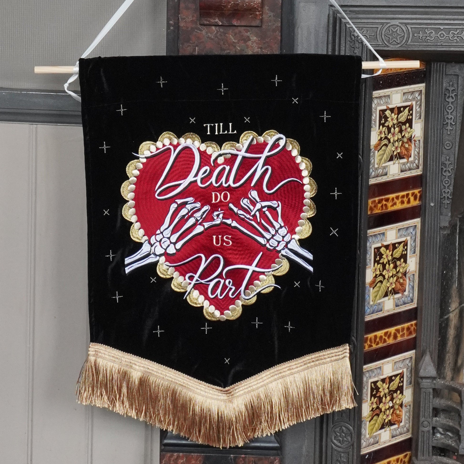 A black hanging banner with gold fringe and a red heart design reading 'Till Death Do Us Part', suspended in front of a fireplace in an indoor room.