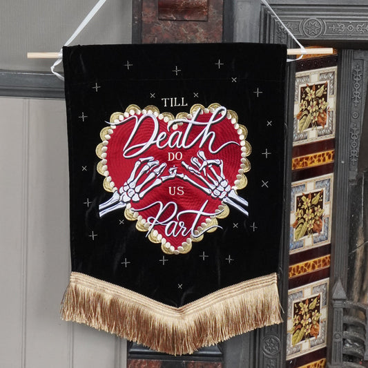 A black hanging banner with gold fringe and a red heart design reading 'Till Death Do Us Part', suspended in front of a fireplace in an indoor room.