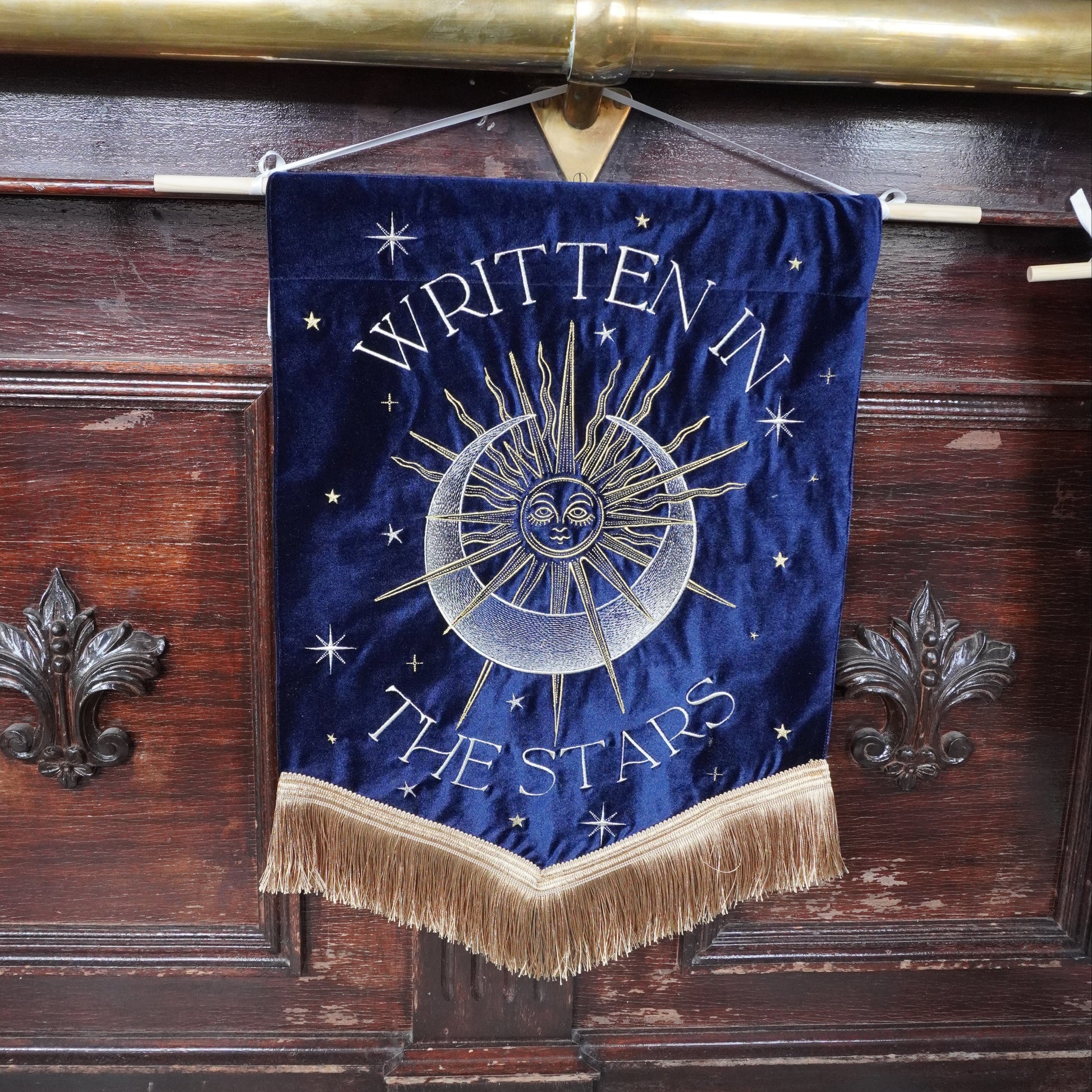 A dark blue velvet banner with gold fringe and a central sun-and-moon emblem, hanging on a wooden bar counter in a pub. The banner reads 'WRITTEN IN THE STARS' in gold lettering.