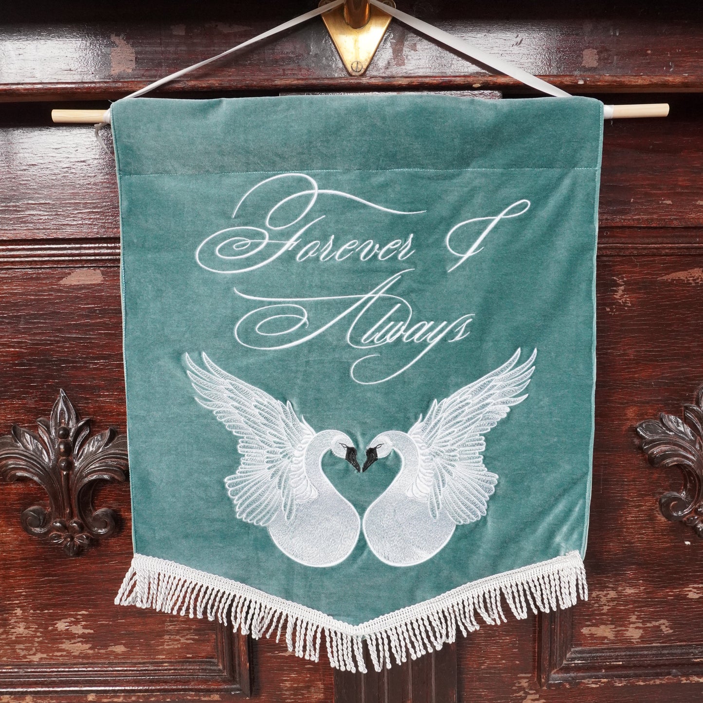 A teal velvet hanging banner with white fringe, featuring two white swans forming a heart shape and the words 'Forever & Always' in white script, displayed against a dark wooden cabinet.