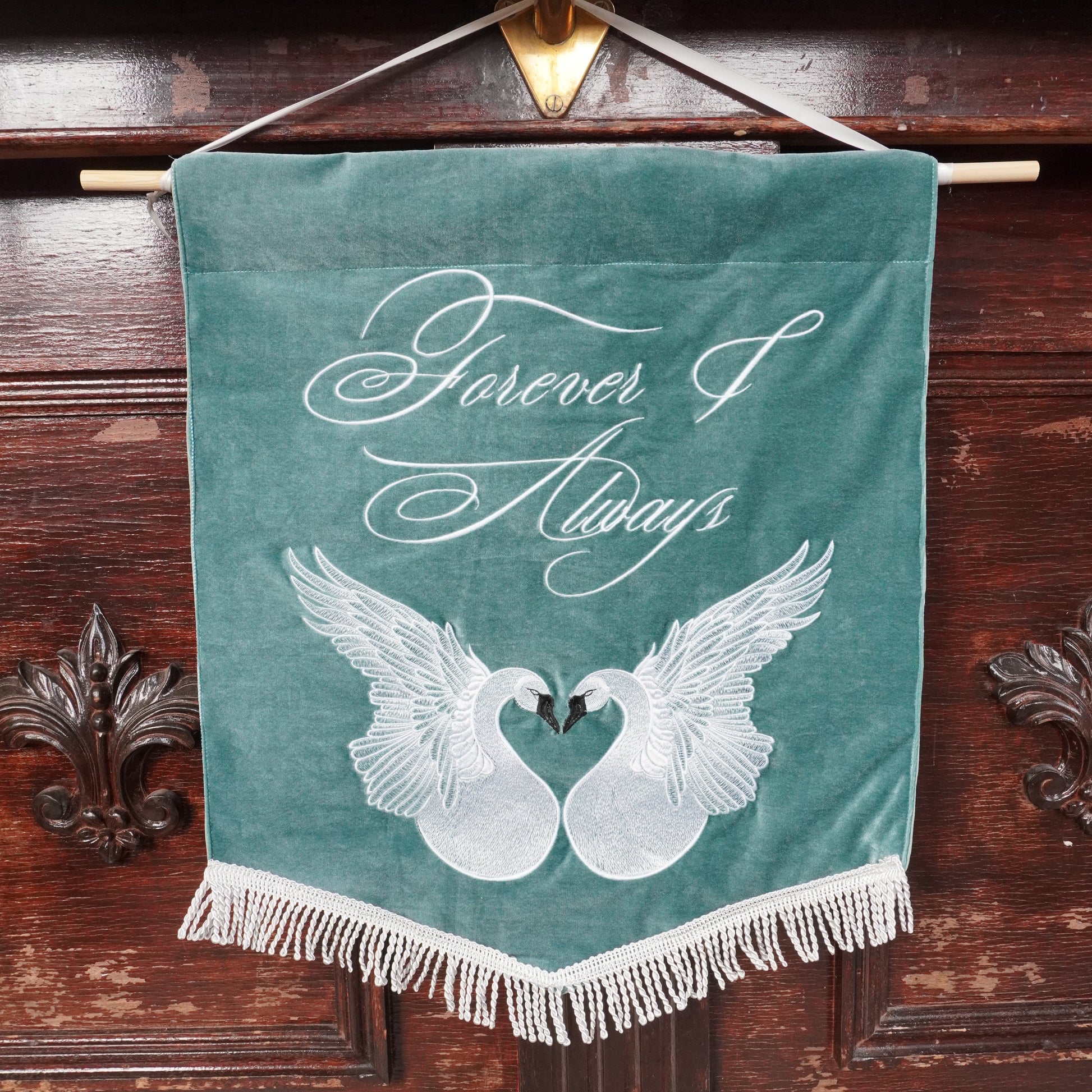 A teal velvet hanging banner with white fringe, featuring two white swans forming a heart shape and the words 'Forever & Always' in white script, displayed against a dark wooden cabinet.
