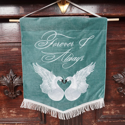 A teal velvet hanging banner with white fringe, featuring two white swans forming a heart shape and the words 'Forever & Always' in white script, displayed against a dark wooden cabinet.