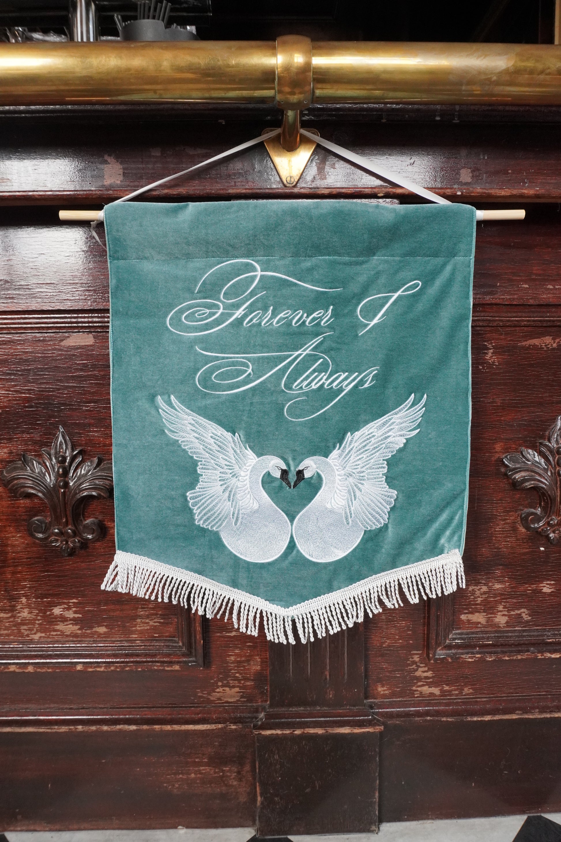 A teal velvet hanging banner with white fringe, featuring the words 'Forever & Always' and two white swans facing each other. The banner hangs from a brass rod against a dark wooden cabinet.