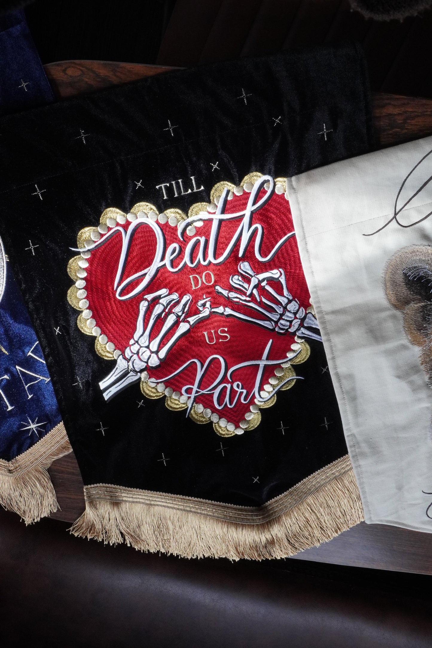 A close-up of a decorative embroidered textile featuring a black velvet-like background with a large central red heart design outlined in gold, white skeleton hands, and the words 'Death Before Us All.' A cream fringed border runs along the right side, and part of a white fabric section with a dog illustration is visible at the top.