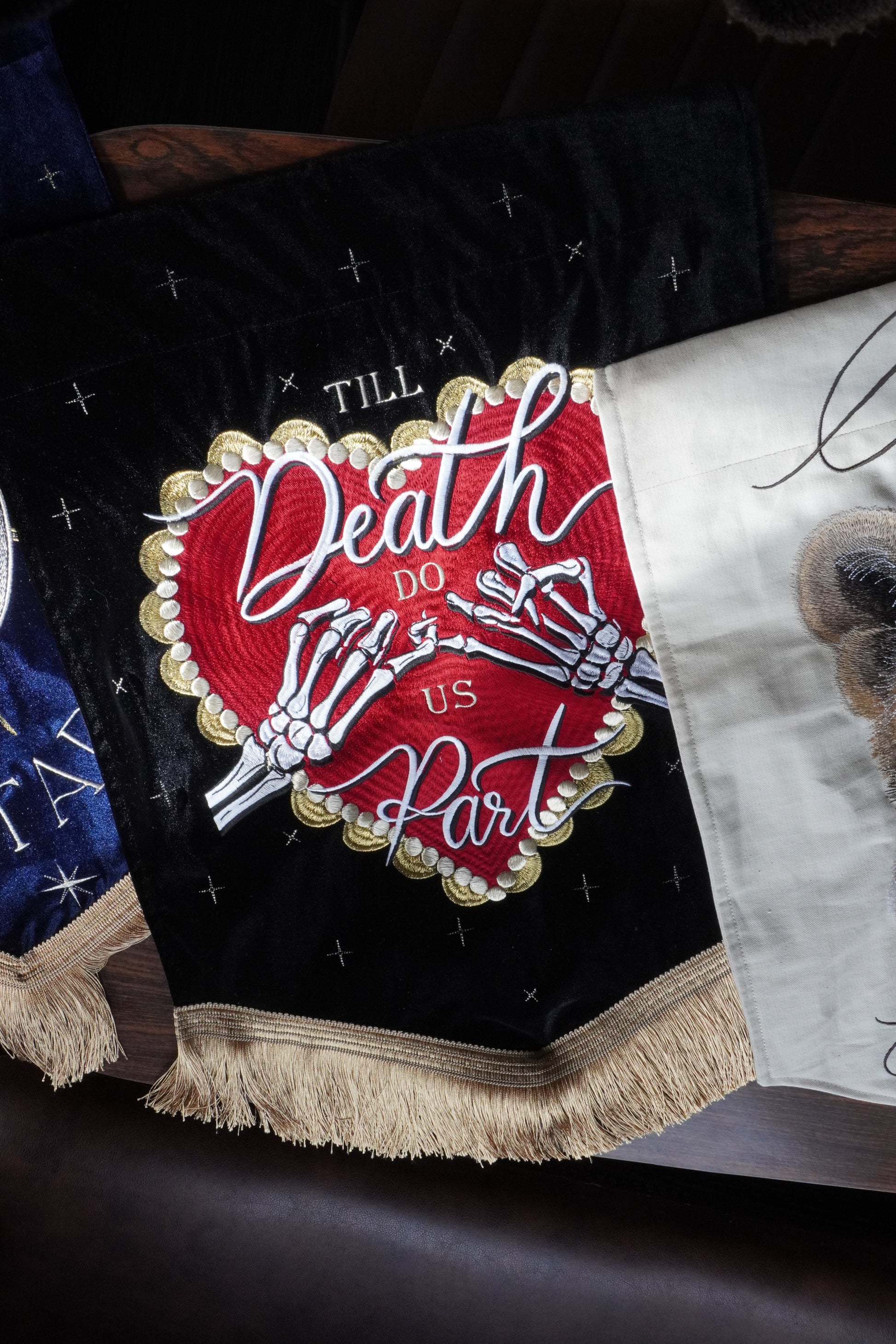 A close-up of a decorative embroidered textile featuring a black velvet-like background with a large central red heart design outlined in gold, white skeleton hands, and the words 'Death Before Us All.' A cream fringed border runs along the right side, and part of a white fabric section with a dog illustration is visible at the top.