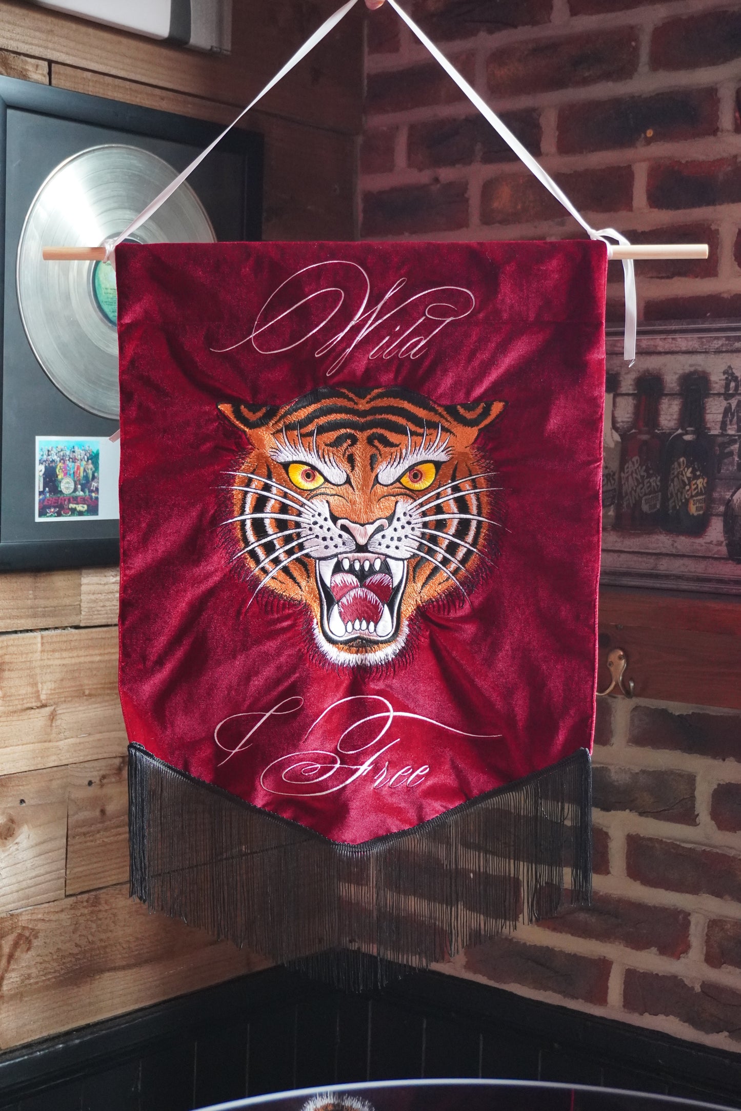 A red velvet wall banner featuring a fierce tiger face illustration and the words 'Wild' and 'Free' in white script, hanging from a wooden rod in a rustic indoor setting with brick and wood walls.