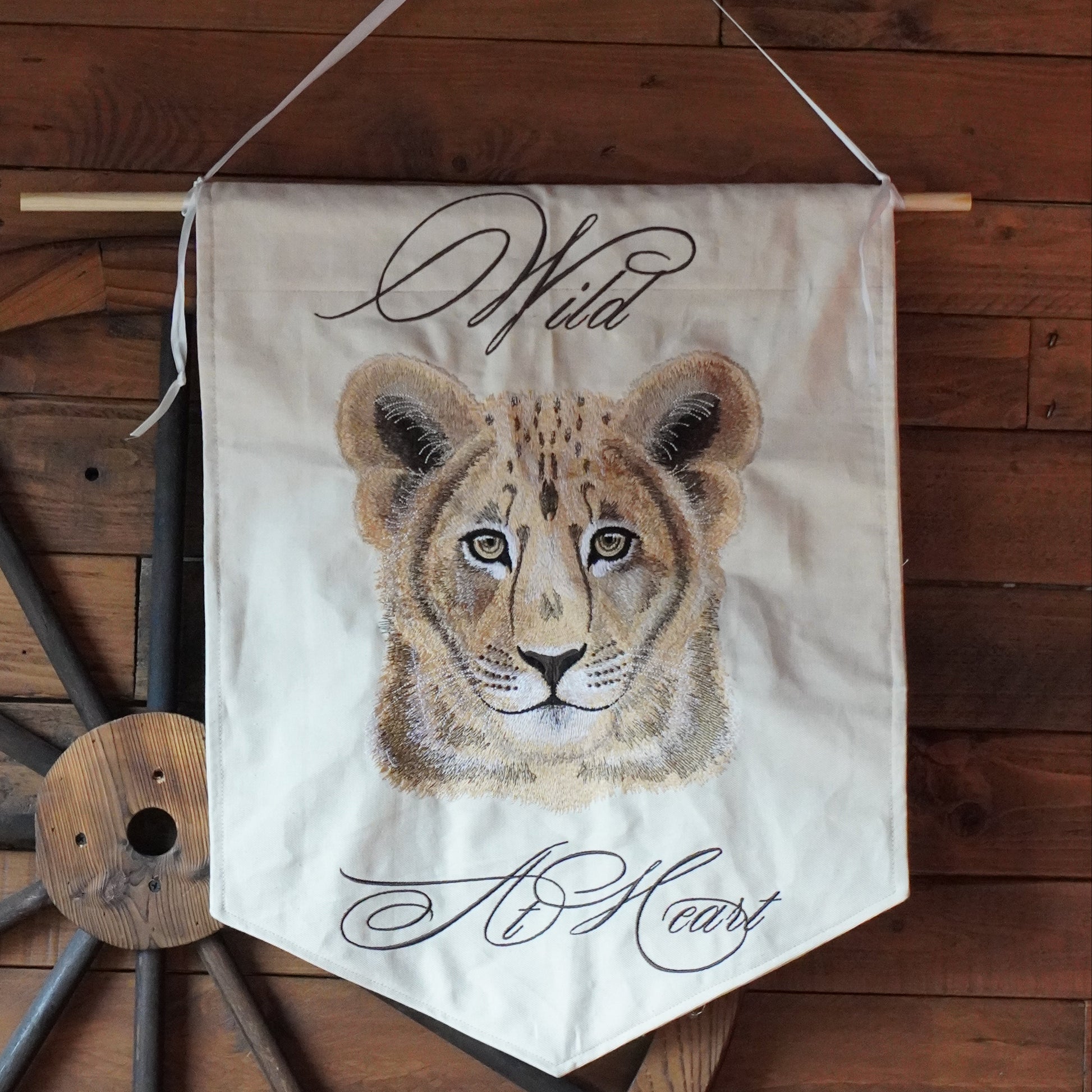 A cream-colored fabric wall banner featuring an embroidered cheetah cub design, with the words 'Wild Heart' in black script, hanging from a wooden dowel against a rustic dark wood plank wall.