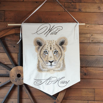 A cream-colored fabric pennant banner featuring a painted cheetah cub face with the words 'Wild' at the top and 'No Heart' at the bottom, hanging from a wooden dowel against a rustic wood plank wall with a large wooden wagon wheel prop.