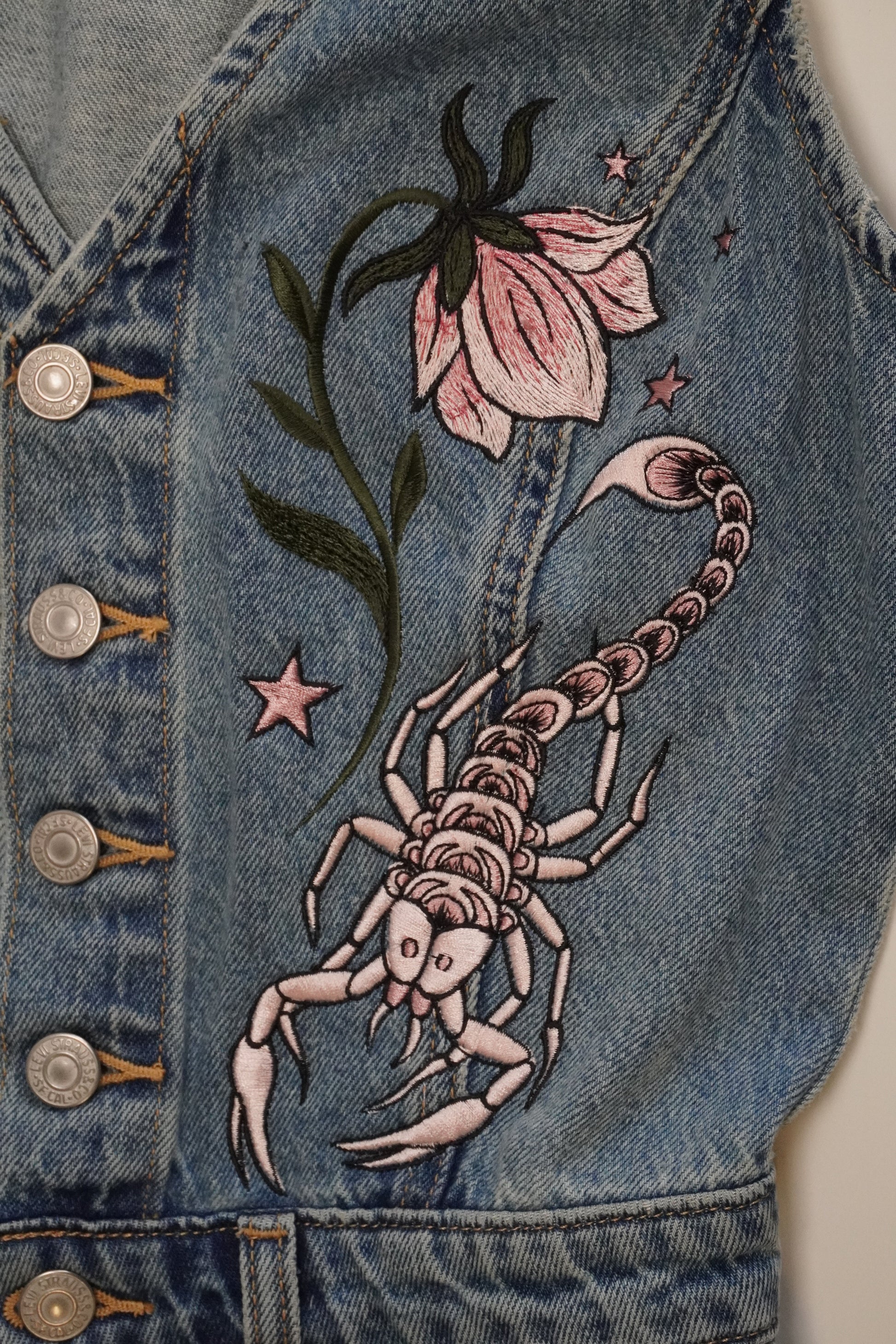 Close-up of a blue denim garment featuring embroidered details of a scorpion, a pink flower, and small stars, with metal snap buttons visible along the top edge.